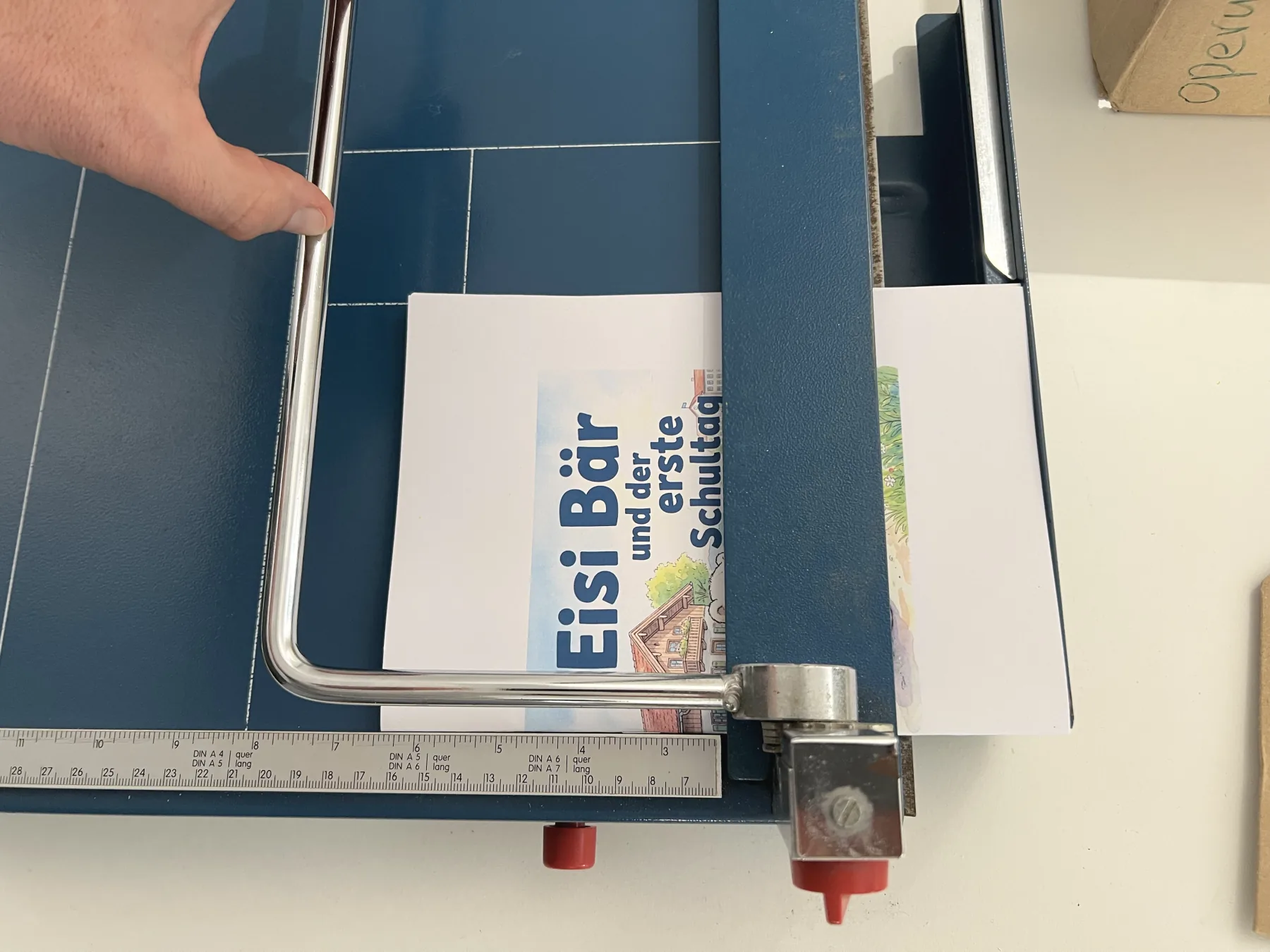 Das gefaltete Büchlein liegt in einer Schneidemaschine, der Hebel ist über die offene Kante gelegt.